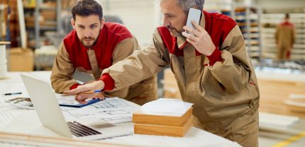 Mid adult worker communicating over mobile phone while using laptop with his colleagues at carpentry workshop.