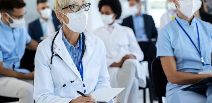 Mature female doctor wearing protective face mask and writing notes while attending educational event at conference hall.